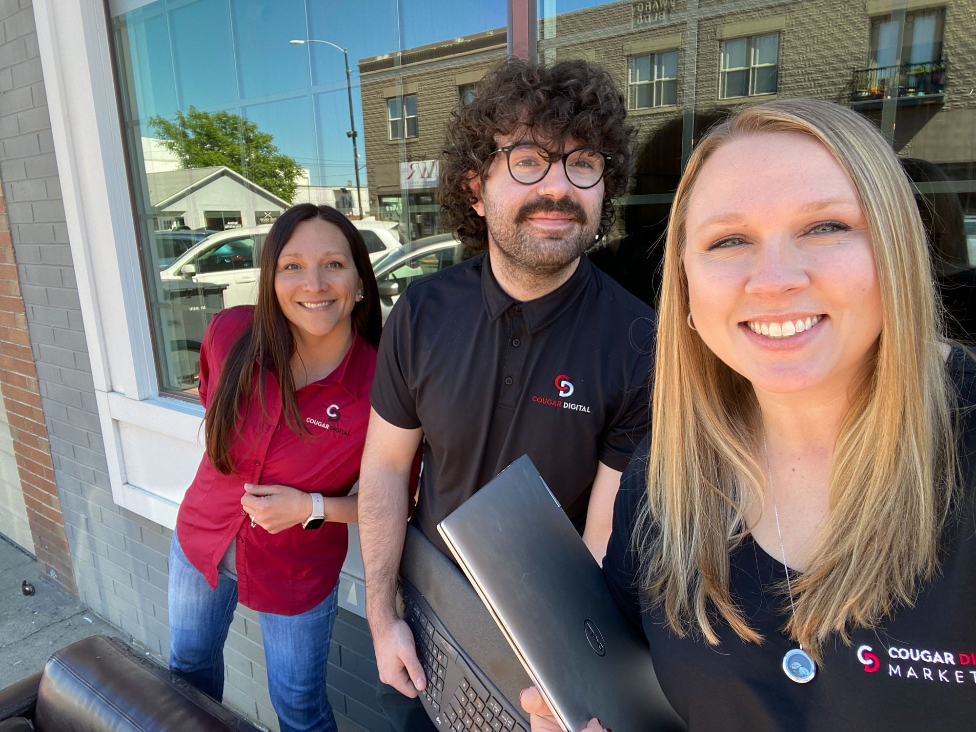 Three smiling marketers outdoors with laptops.