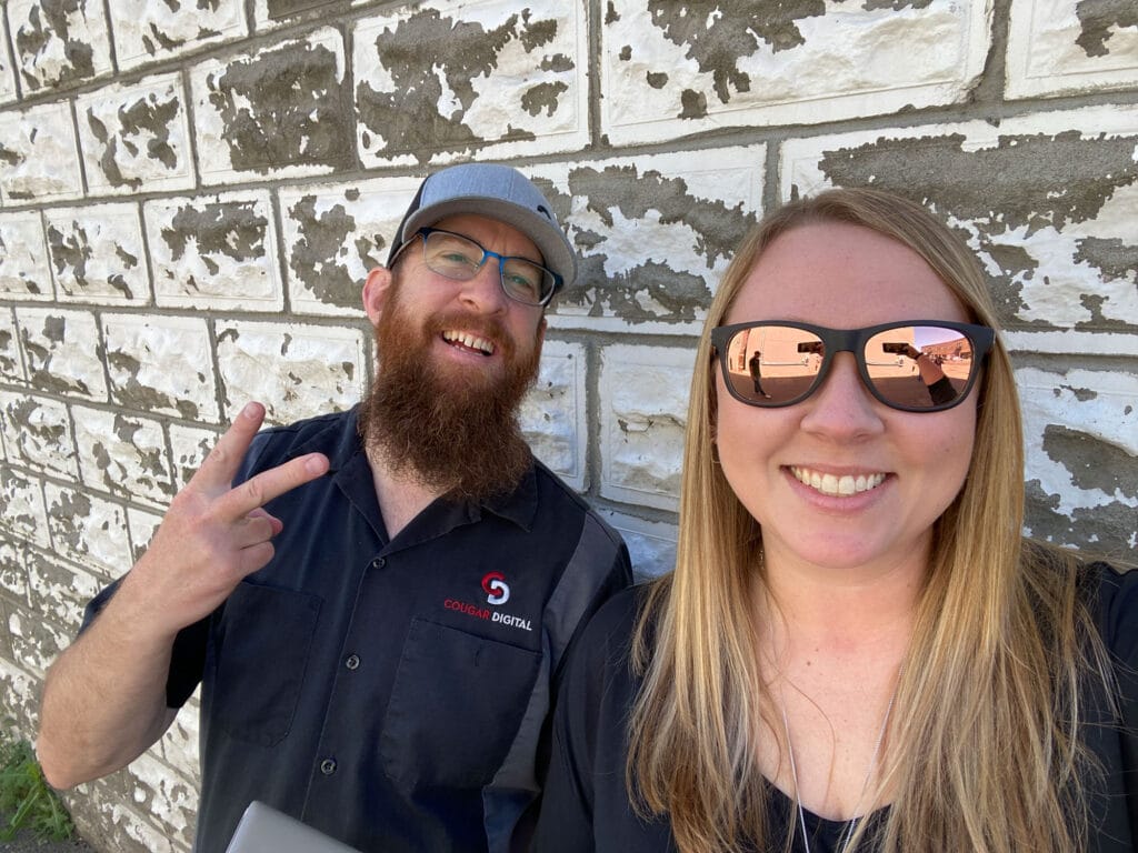 Two people smiling against a brick wall backdrop.