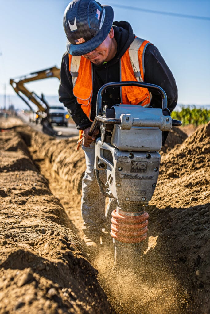 Construction worker operating a compactor on dirt trench.