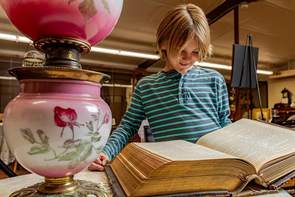 Child reading large book beside vintage lamp.