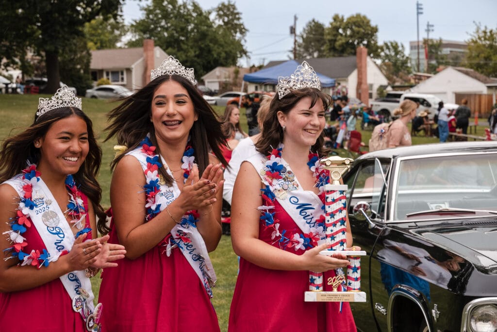 Three girls in crowns, holding trophy at outdoor event.