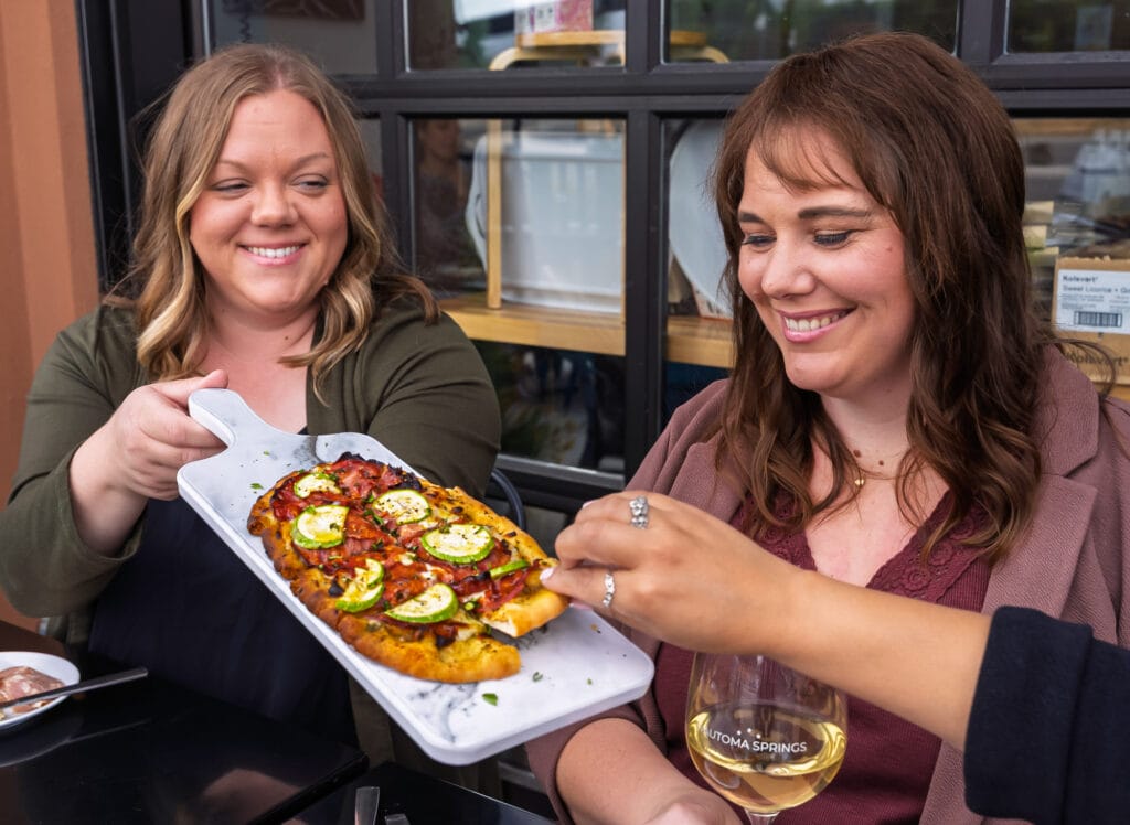 Two women sharing pizza and wine, smiling.